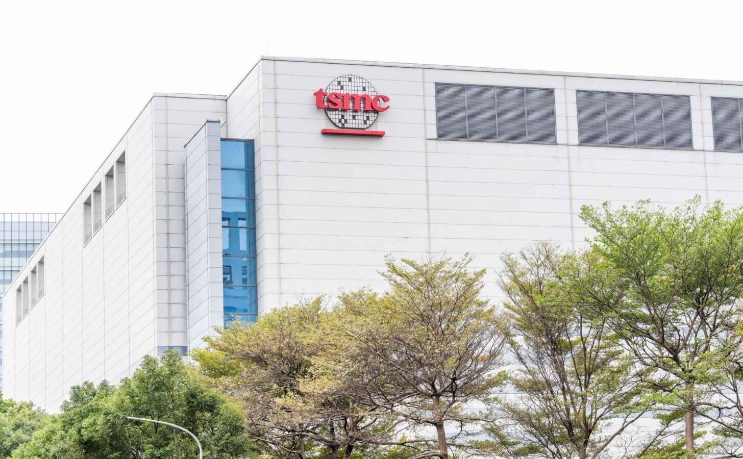 نمای بیرونی ساختمان مدرن کارخانه تولید نیمههادی شرکت TSMC با نمای سفید و پنجرههای شیشهای آبیرنگ، با لوگوی قرمز TSMC در بالای سازه و ردیفی از درختان سرسبز در پیشزمینه.