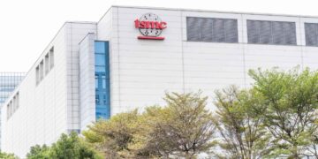 نمای بیرونی ساختمان مدرن کارخانه تولید نیمههادی شرکت TSMC با نمای سفید و پنجرههای شیشهای آبیرنگ، با لوگوی قرمز TSMC در بالای سازه و ردیفی از درختان سرسبز در پیشزمینه.
