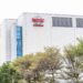 نمای بیرونی ساختمان مدرن کارخانه تولید نیمههادی شرکت TSMC با نمای سفید و پنجرههای شیشهای آبیرنگ، با لوگوی قرمز TSMC در بالای سازه و ردیفی از درختان سرسبز در پیشزمینه.