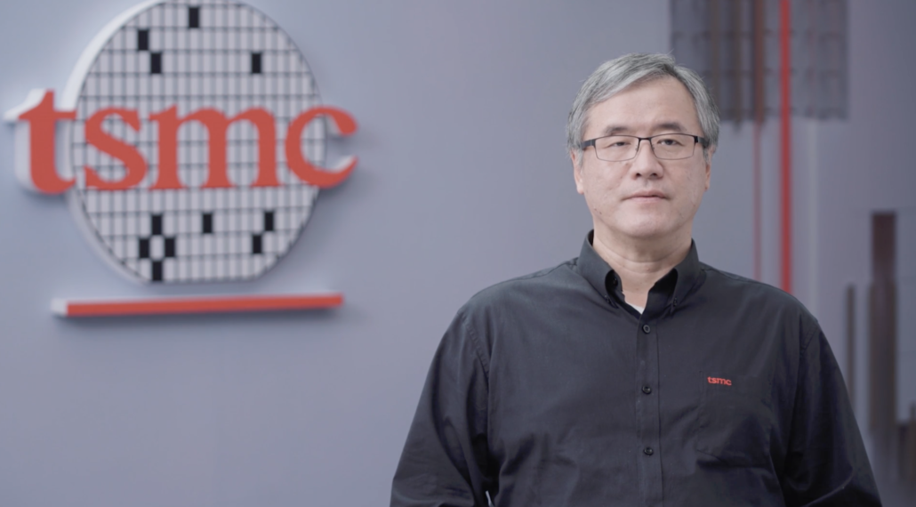 پرتره وای جی می (Y.J. Mii)، معاون ارشد توسعه فناوری و تحقیق و توسعه در TSMC، با پیراهن مشکی و عینک، در حال ارائه سخنرانی مقابل دیوار رسمی خاکستری با لوگوی برجسته قرمز و دیسک سیلیکونی نمادین TSMC.