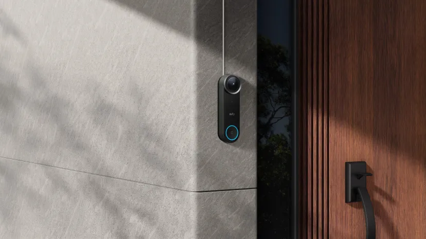 زنگ ویدیویی هوشمند یوفی مدل Video Doorbell S4 با کلید دارای نور آبی، نصبشده روی دیوار سنگی در کنار درب ورودی چوبی خانه.