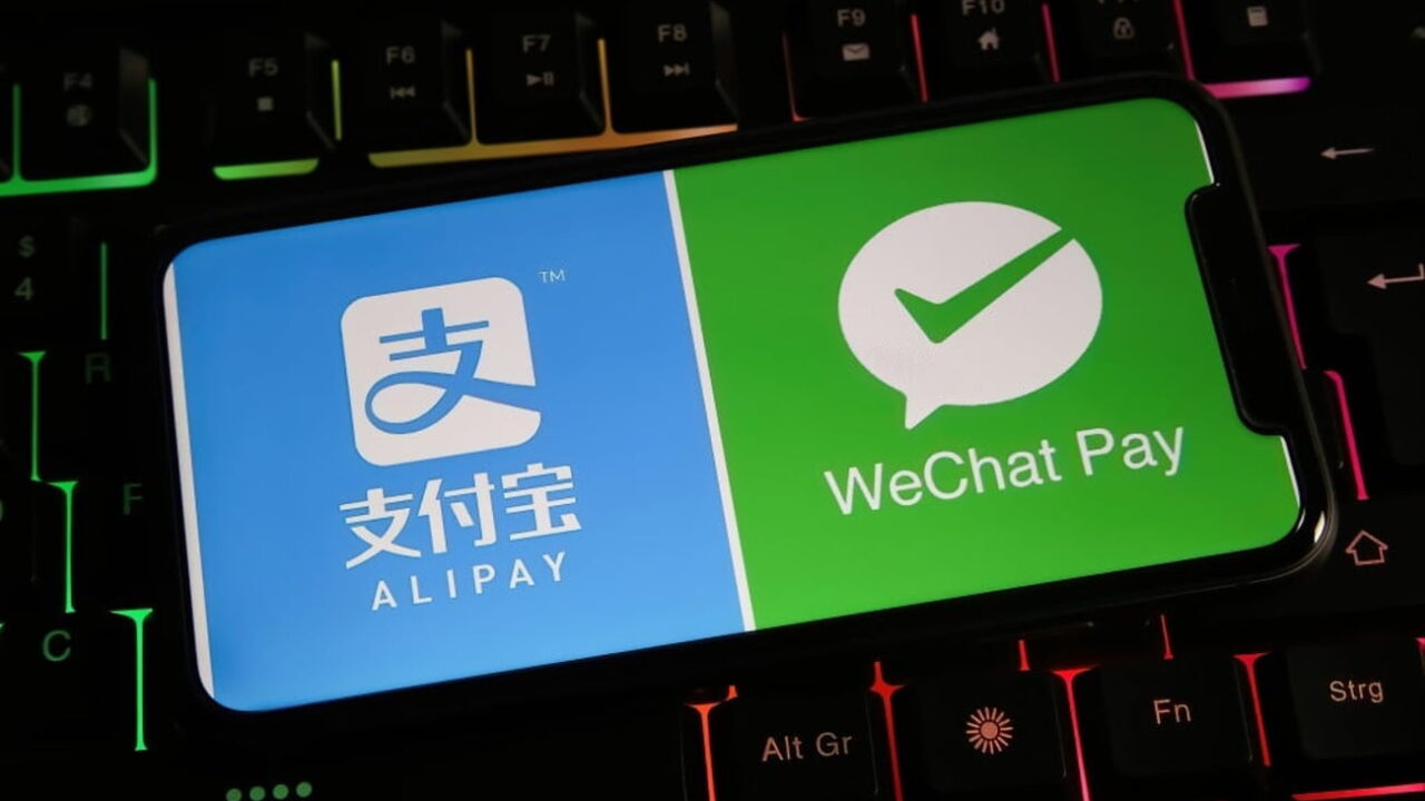یک گوشی هوشمند روی صفحهکلید گیمینگ با نور پسزمینه رنگی که لوگوهای Alipay و WeChat Pay را روی نمایشگر خود نشان میدهد.