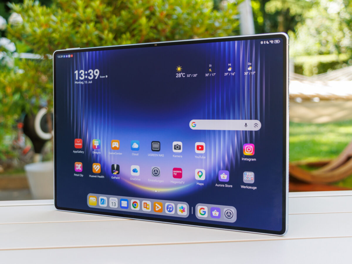 نمای روبهرو تبلت هواوی MatePad Pro Max با نمایشگر بزرگ، رابط کاربری HarmonyOS، صفحه اصلی شامل ساعت ۱۳:۳۹، ابزارک هوا و آیکونهای اپلیکیشنهایی مانند یوتیوب و اینستاگرام، روی میز چوبی سفید در فضای باز.