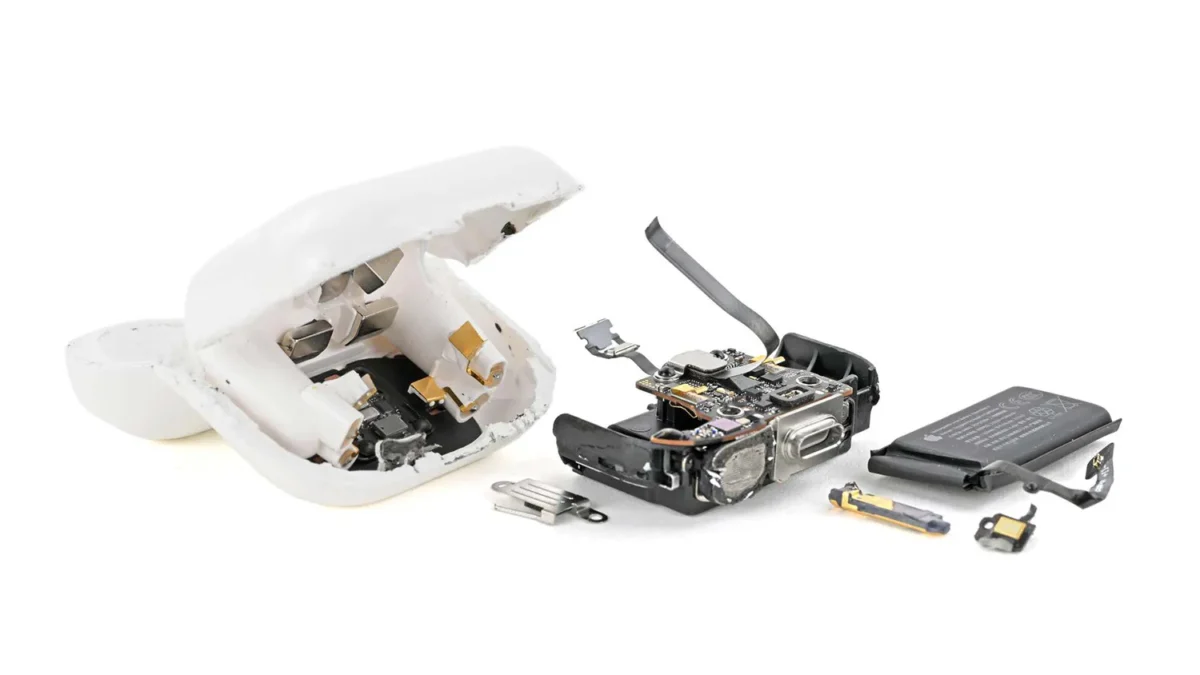 عکس جداسازی قطعات (teardown) جعبه شارژ AirPods 5 توسط iFixit روی پسزمینه سفید. پوسته سفید جعبه شکافته شده و قطعات داخلی شامل مگنتها و پینهای شارژ، برد الکترونیکی با درگاه USB-C و کابل فلکسی بلند، باتری مشکی مستطیلی و چند قطعه فلزی کوچک کنار هم چیده شدهاند.
