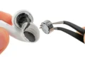 عکس کلوزآپ از جداسازی قطعات AirPods 5. دستی یک ایرباد سفید با پوسته شکافته را نگه داشته و با پنس مشکی، درایور بلندگوی نقره‌ای کوچک را از داخل آن بیرون می‌کشد. پس‌زمینه تصویر کاملاً سفید است.