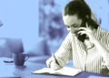 زن جوان در حال صحبت با گوشی موبایل و یادداشتبرداری با خودکار در دفترچه روی میز کار