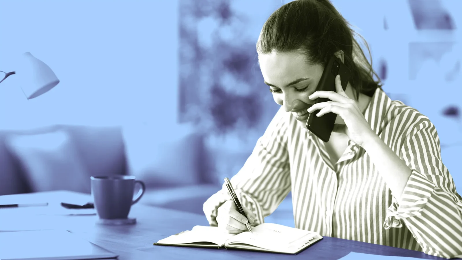 زن جوان در حال صحبت با گوشی موبایل و یادداشتبرداری با خودکار در دفترچه روی میز کار