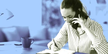 زن جوان در حال صحبت با گوشی موبایل و یادداشتبرداری با خودکار در دفترچه روی میز کار