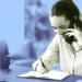 زن جوان در حال صحبت با گوشی موبایل و یادداشت‌برداری با خودکار در دفترچه روی میز کار