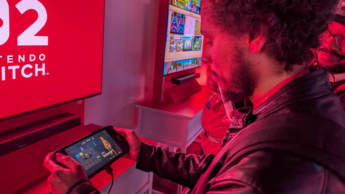 شرکتکنندهای در یک رویداد عمومی با نورپردازی قرمز، در حال امتحان کردن Nintendo Switch 2 در حالت دستی است که صفحهنمایشی رنگی از منوی بازی را نشان میدهد و بنر بزرگ برند در پسزمینه دیده میشود.