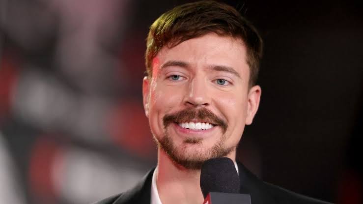 نمای نزدیک از چهره مستربیست (MrBeast) در حال لبخند زدن با کت و پیراهن رسمی در دست داشتن میکروفون حین مصاحبه.
