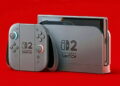 رندر کنسول Nintendo Switch 2 با کنترلرهای Joy-Con رنگی روی پسزمینه قرمز پررنگ که طراحی جدید و تغییریافته دستگاه را در مقایسه با نسل قبلی به نمایش میگذارد.