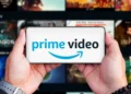 دو دست یک گوشی موبایل را به صورت افقی گرفتهاند که لوگوی آبی و مشکی Prime Video با آیکون لبخند آمازون روی نمایشگر آن درج شده است؛ پسزمینه تار، شبکهای از کاورهای محتوای ویدئویی سرویس استریم را نشان میدهد.