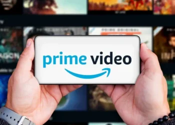 دو دست یک گوشی موبایل را به صورت افقی گرفته‌اند که لوگوی آبی و مشکی Prime Video با آیکون لبخند آمازون روی نمایشگر آن درج شده است؛ پس‌زمینه تار، شبکه‌ای از کاورهای محتوای ویدئویی سرویس استریم را نشان می‌دهد.