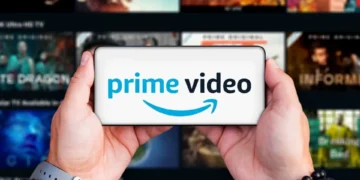 دو دست یک گوشی موبایل را به صورت افقی گرفتهاند که لوگوی آبی و مشکی Prime Video با آیکون لبخند آمازون روی نمایشگر آن درج شده است؛ پسزمینه تار، شبکهای از کاورهای محتوای ویدئویی سرویس استریم را نشان میدهد.