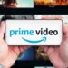 دو دست یک گوشی موبایل را به صورت افقی گرفته‌اند که لوگوی آبی و مشکی Prime Video با آیکون لبخند آمازون روی نمایشگر آن درج شده است؛ پس‌زمینه تار، شبکه‌ای از کاورهای محتوای ویدئویی سرویس استریم را نشان می‌دهد.