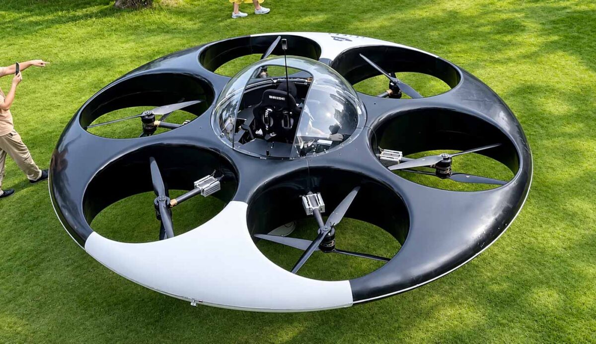 نمای نزدیک از بشقاب پرنده eVTOL با بدنه دایره‌ای و شش مجموعه پروانه در محوطه‌ای سبز