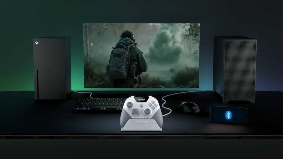 میز گیمینگ تیره با کنترلر سفید خاکستری SteelSeries Aeon Pro روی پایه شارژ اختصاصی در مرکز؛ کنسول مشکی Xbox Series X با نور سبز در سمت چپ، کیس کامپیوتر مینی در سمت راست، مانیتور در حال نمایش بازی جنگی شوتر اولشخص، کیبورد مکانیکی، ماوس گیمینگ و گوشی نمایشدهنده لوگوی بلوتوث.