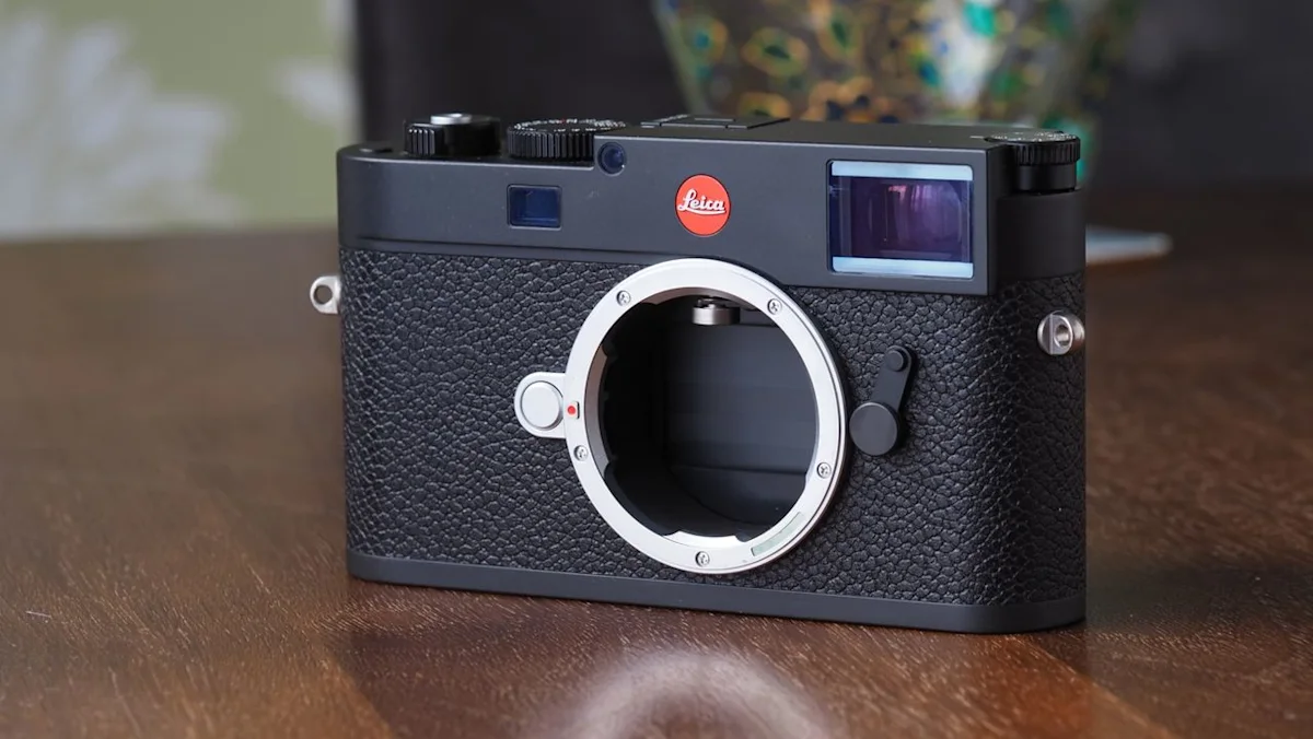 دوربین عکاسی دیجیتال لایکا (Leica) با بدنهی مشکی چرمی و مانت لنز فلزی بدون لنز، قرارگرفته روی سطح چوبی.