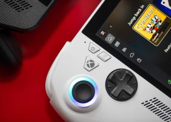 عکس نزدیک کنسول دستی ROG Xbox Ally: نمای نزدیک از بخش سمت چپ کنسول دستی سفیدرنگ ROG Xbox Ally X روی میز قرمزرنگ که آنالوگ روشن با حلقه نورانی آبی، دی‌پد مشکی، دکمه Xbox و دکمه‌های سفارشی ASUS و صفحه نمایش آن با رابط Xbox و کاور بازی Cuphead و برچسب Game Pass دیده می‌شود.