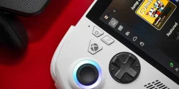 عکس نزدیک کنسول دستی ROG Xbox Ally: نمای نزدیک از بخش سمت چپ کنسول دستی سفیدرنگ ROG Xbox Ally X روی میز قرمزرنگ که آنالوگ روشن با حلقه نورانی آبی، دیپد مشکی، دکمه Xbox و دکمههای سفارشی ASUS و صفحه نمایش آن با رابط Xbox و کاور بازی Cuphead و برچسب Game Pass دیده میشود.