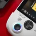 عکس نزدیک کنسول دستی ROG Xbox Ally: نمای نزدیک از بخش سمت چپ کنسول دستی سفیدرنگ ROG Xbox Ally X روی میز قرمزرنگ که آنالوگ روشن با حلقه نورانی آبی، دیپد مشکی، دکمه Xbox و دکمههای سفارشی ASUS و صفحه نمایش آن با رابط Xbox و کاور بازی Cuphead و برچسب Game Pass دیده میشود.