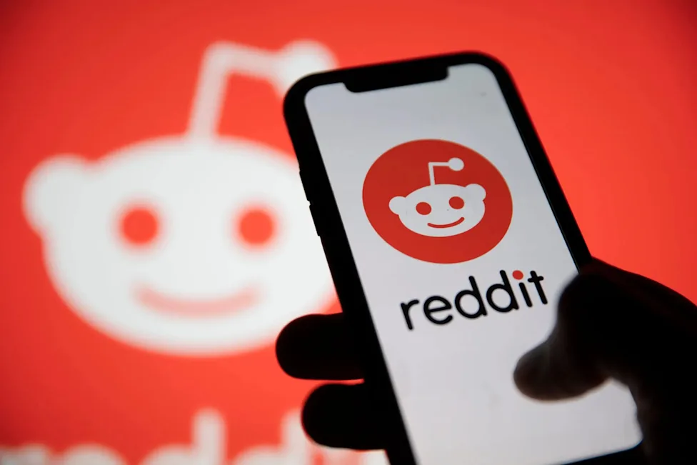 دستی که یک گوشی هوشمند را با نمایشگر بازشده روی لوگو و نام ردیت (Reddit) نگه داشته، در حالی که پس‌زمینه قرمز با لوگوی بزرگ ردیت دیده می‌شود.