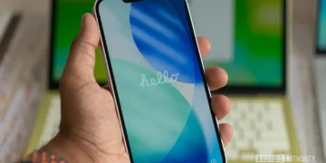 دستی گوشی آیفون را در حال بوت و نمایش صفحه «hello» گرفته؛ در پس‌زمینه لپ‌تاپی سبز رنگ دیده می‌شود.