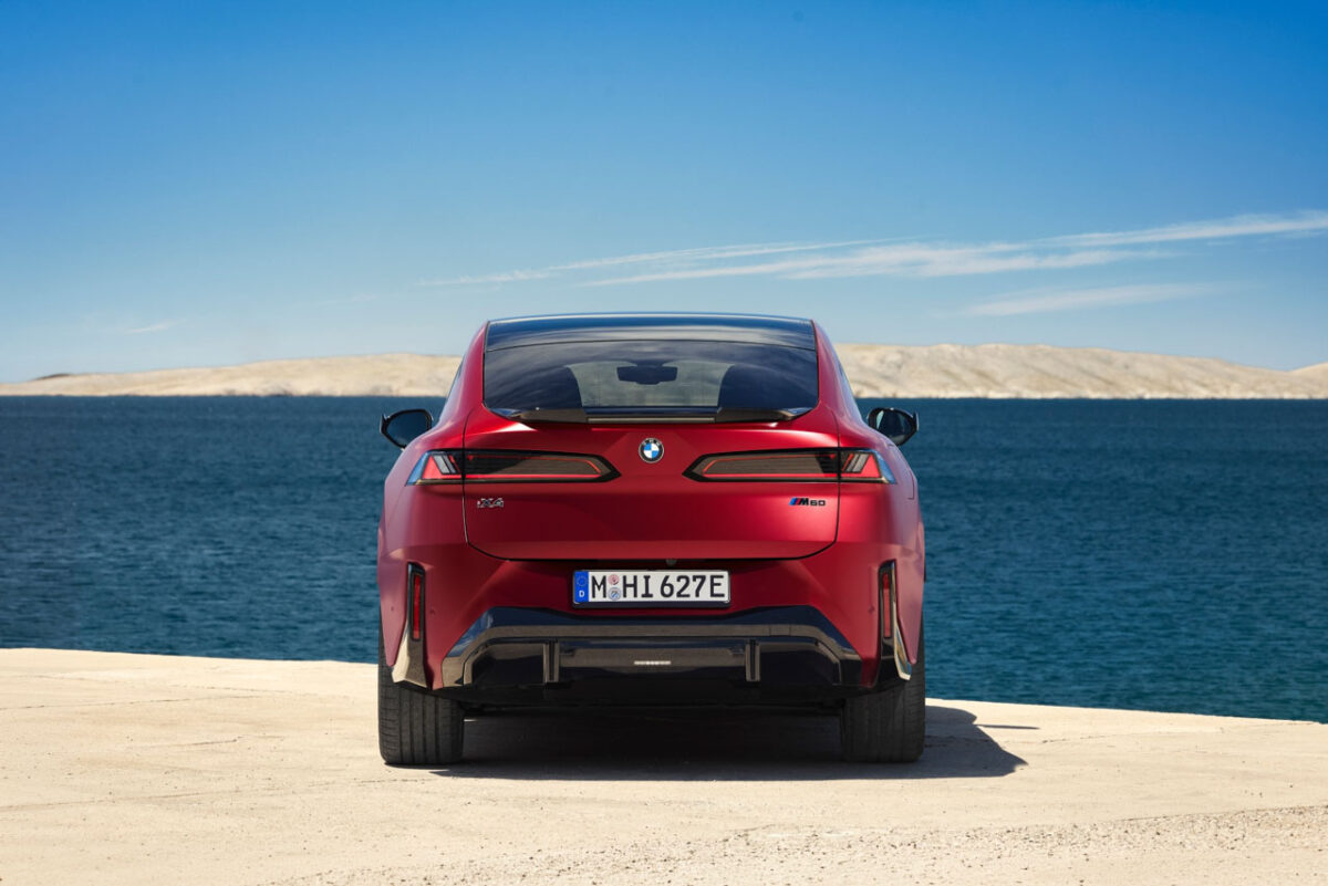 نمای مستقیم عقب شاسیبلند برقی BMW iX4 M60 با چراغهای افقی باریک، نشان BMW و سپر اسپرت در کنار دریا