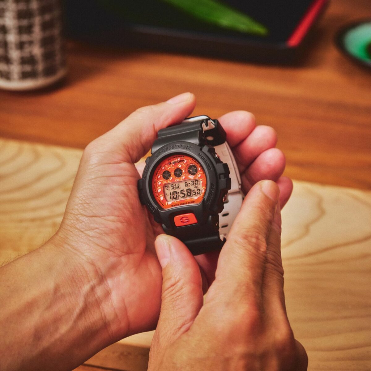 نمای نزدیک از دستان یک فرد که ساعت مچی دیجیتال کاسیو جیشاک مشکی مدل DW-6900 با صفحهی طرح خاویار نارنجی را روی یک میز چوبی در دست نگه داشته است.