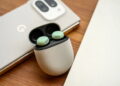 نمای نزدیک محصول از کیس شارژ سفید رنگ بادزن‌های گوگل Pixel Buds در حالت باز روی میز چوبی، با دو ایرباد سبز داخل کیس و پشت‌بند گوشی موبایل گوگل با دوربین بزرگ در پس‌زمینه‌ی محو
