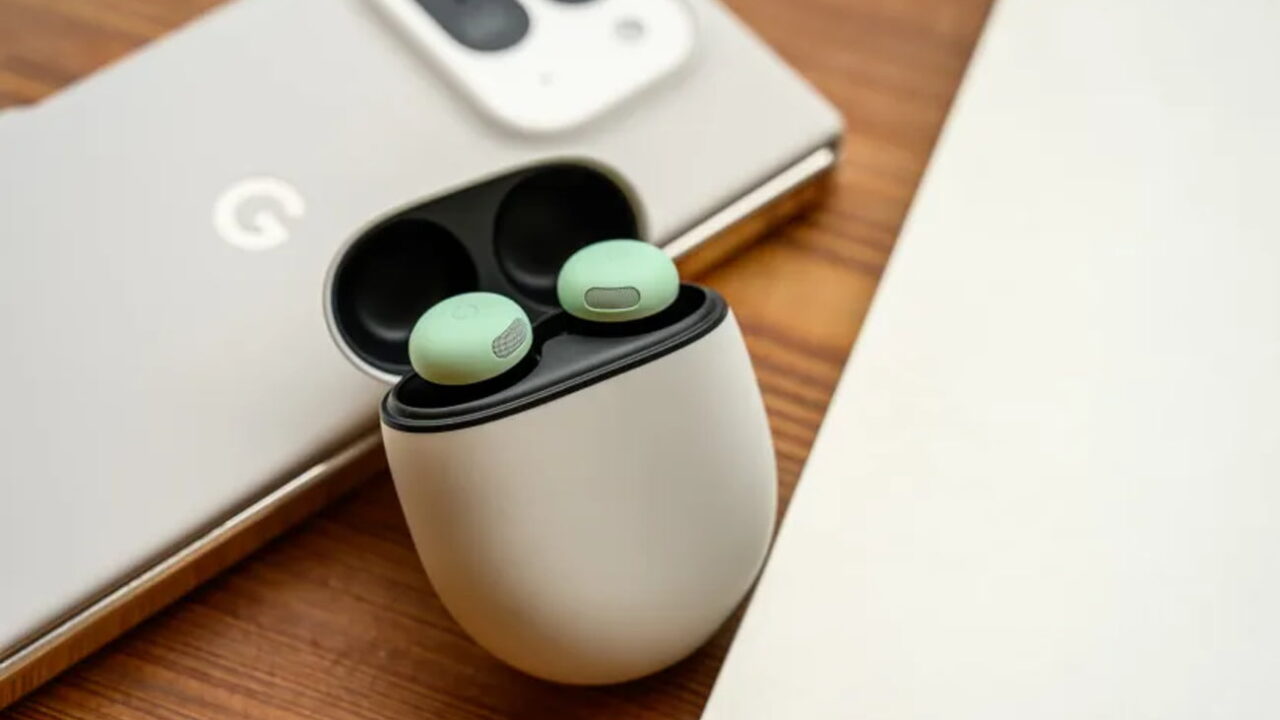نمای نزدیک محصول از کیس شارژ سفید رنگ بادزن‌های گوگل Pixel Buds در حالت باز روی میز چوبی، با دو ایرباد سبز داخل کیس و پشت‌بند گوشی موبایل گوگل با دوربین بزرگ در پس‌زمینه‌ی محو