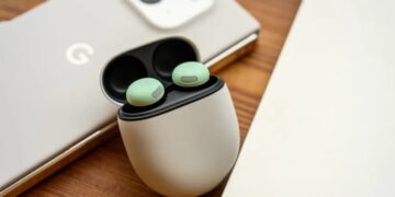 نمای نزدیک محصول از کیس شارژ سفید رنگ بادزن‌های گوگل Pixel Buds در حالت باز روی میز چوبی، با دو ایرباد سبز داخل کیس و پشت‌بند گوشی موبایل گوگل با دوربین بزرگ در پس‌زمینه‌ی محو