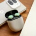 نمای نزدیک محصول از کیس شارژ سفید رنگ بادزنهای گوگل Pixel Buds در حالت باز روی میز چوبی، با دو ایرباد سبز داخل کیس و پشتبند گوشی موبایل گوگل با دوربین بزرگ در پسزمینهی محو