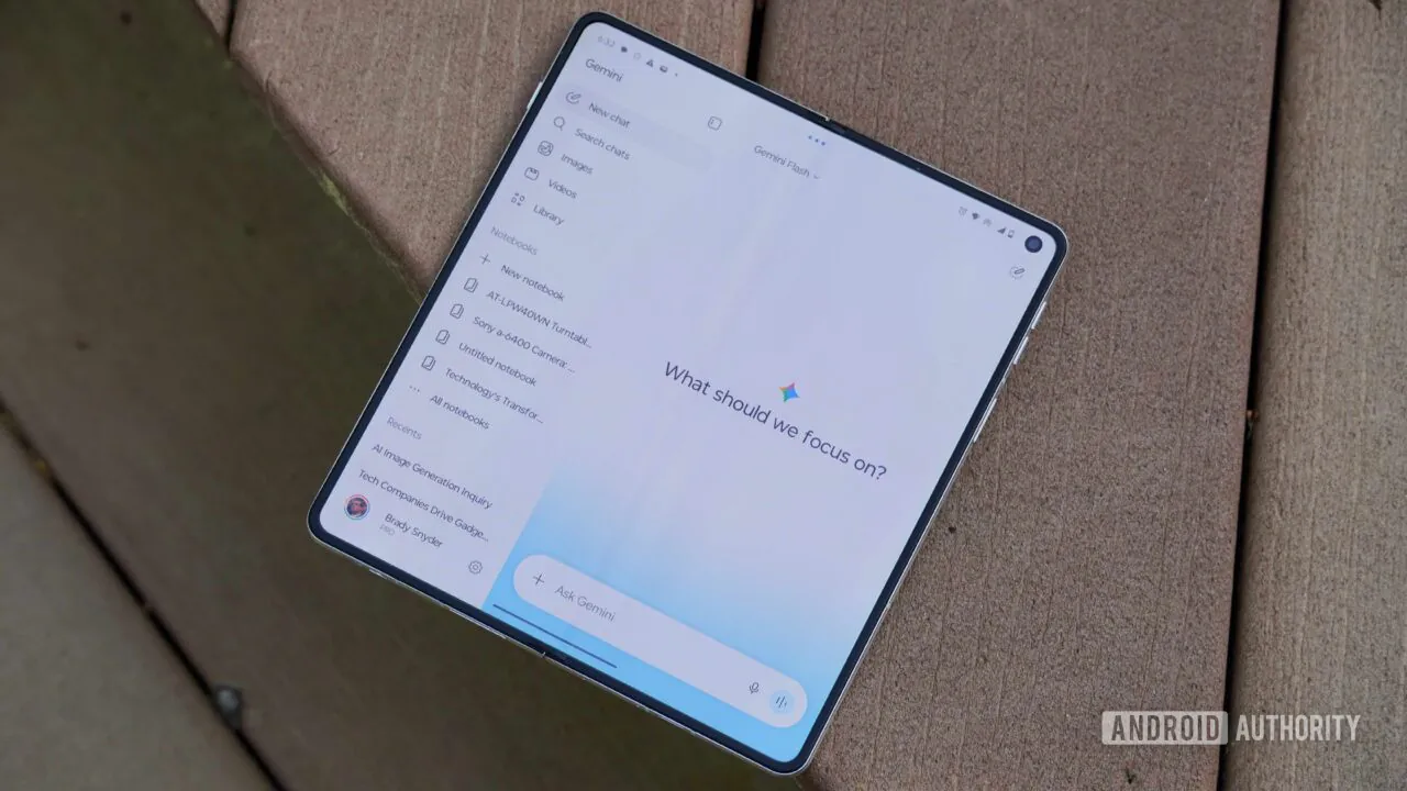 یک گوشی هوشمند تاشو (Foldable) باز شده روی سطح چوبی قرار دارد که رابط کاربری اپلیکیشن جمینای شامل منوی کناری یادداشتها و عبارت «What should we focus on?» را نمایش میدهد