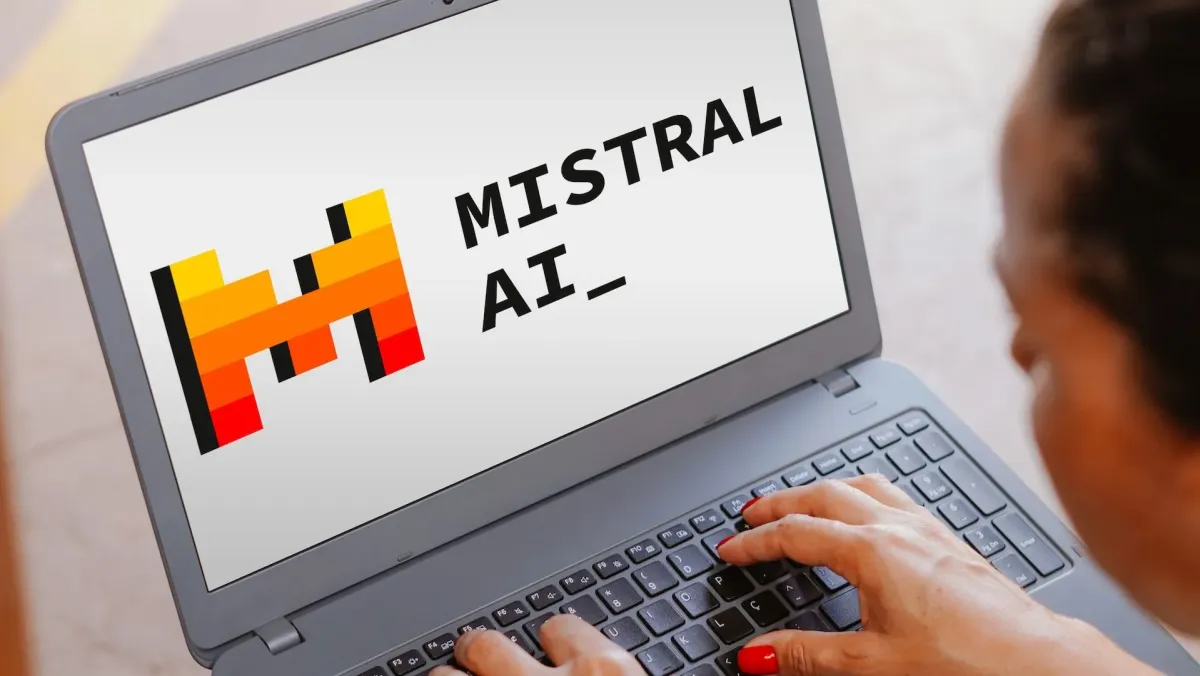 زنی با ناخنهای لاکقرمز در حال تایپ روی لپتاپی است که صفحهنمایش آن لوگوی پیکسلی نارنجی و زرد و نام Mistral AI را نشان میدهد