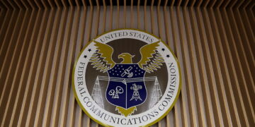 نشان دایرهای شکل کمیسیون فدرال ارتباطات آمریکا (FCC) بر دیوار چوبی نواری نصب شده است. در مرکز نشان، عقابی با بالهای باز و سپری آبیرنگ با نمادهای ارتباطی مانند دکل و ماهواره دیده میشود و عبارت United States Federal Communications Commission در حاشیه دایره نوشته شده است.