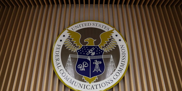 نشان دایره‌ای شکل کمیسیون فدرال ارتباطات آمریکا (FCC) بر دیوار چوبی نواری نصب شده است. در مرکز نشان، عقابی با بال‌های باز و سپری آبی‌رنگ با نمادهای ارتباطی مانند دکل و ماهواره دیده می‌شود و عبارت United States Federal Communications Commission در حاشیه دایره نوشته شده است.