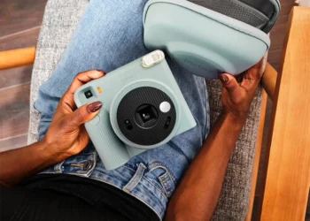 نمای بالای فردی نشسته با شلوار جین آبی که یک دوربین چاپ سریع آبی پاستلی Instax Square SQ2 و کیف محافظ هم‌رنگ آن را روی پای خود نگه داشته است.