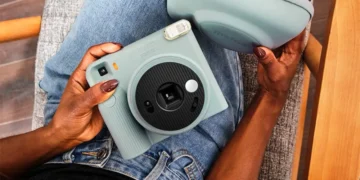 نمای بالای فردی نشسته با شلوار جین آبی که یک دوربین چاپ سریع آبی پاستلی Instax Square SQ2 و کیف محافظ هم‌رنگ آن را روی پای خود نگه داشته است.