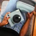 نمای بالای فردی نشسته با شلوار جین آبی که یک دوربین چاپ سریع آبی پاستلی Instax Square SQ2 و کیف محافظ همرنگ آن را روی پای خود نگه داشته است.