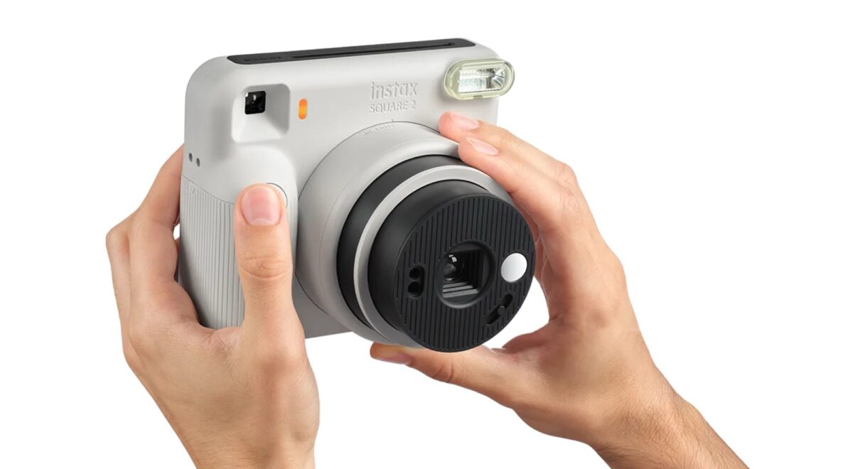 دو دست که یک دوربین چاپ سریع طوسی روشن Instax Square SQ2 را برای عکاسی نگه داشتهاند، با تمرکز بر کلیدها، لنز چرخشی مشکی و فلاش بالایی در پسزمینه سفید.