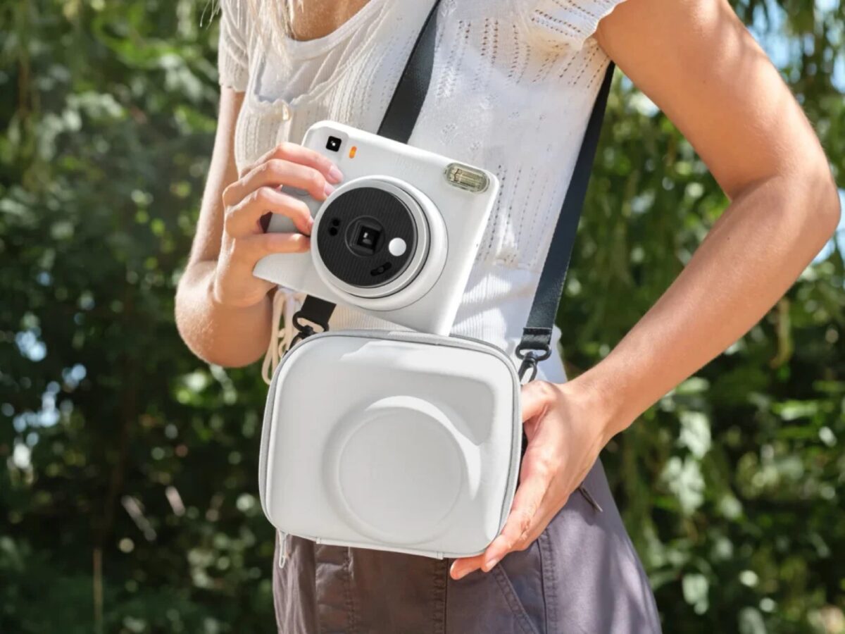 فردی با لباس تابستانی سفید و کیف دوشی سفید، دوربین چاپ سریع سفید Instax Square SQ2 را با بند مشکی روی گردن و دستان خود نگه داشته است.