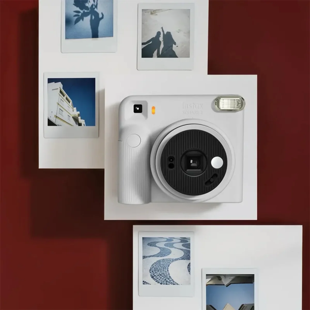 دوربین چاپ سریع سفید رنگ Instax Square SQ2 با لنز مشکی و فلاش نقرهای در مرکز تصویر، احاطهشده توسط چهار عکس چاپشده مربعی روی پسزمینه زرشکی.