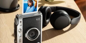 دوربین اینستاکس mini Evo 2 روی میز چوبی با هدفون و چند عکس چاپشده اطراف آن.