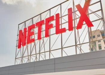 تابلوی بزرگ و قرمز رنگ نام Netflix روی یک سازه فلزی داربستی در فضای باز، زیر آسمانی ابری و در کنار یک ساختمان مسکونی مدرن.