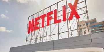 تابلوی بزرگ و قرمز رنگ نام Netflix روی یک سازه فلزی داربستی در فضای باز، زیر آسمانی ابری و در کنار یک ساختمان مسکونی مدرن.