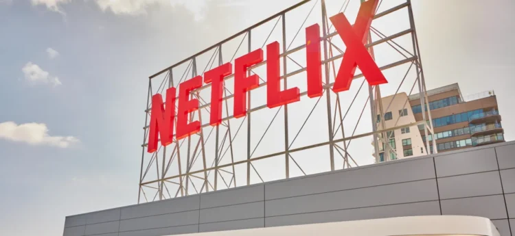 تابلوی بزرگ و قرمز رنگ نام Netflix روی یک سازه فلزی داربستی در فضای باز، زیر آسمانی ابری و در کنار یک ساختمان مسکونی مدرن.