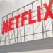 تابلوی بزرگ و قرمز رنگ نام Netflix روی یک سازه فلزی داربستی در فضای باز، زیر آسمانی ابری و در کنار یک ساختمان مسکونی مدرن.