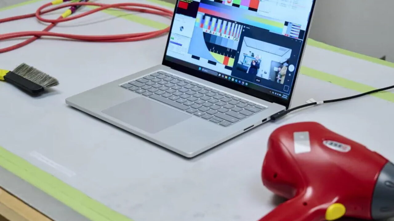 عکس از یک لپتاپ Surface نقرهای در حالت باز روی میز تست که صفحهنمایش رنگبندیهای نوار رنگی آزمایشی و نرمافزار اندازهگیری رنگ و یک پنجرهی وبکم با حضور افراد را نشان میدهد. در سمت چپ میز، یک برس تمیزکردن گرد و غبار مشکی و سیمهای قرمز و زرد و در سمت راست، یک سشوار صنعتی قرمز با سیمهای مشکی و زرد قرار گرفته است